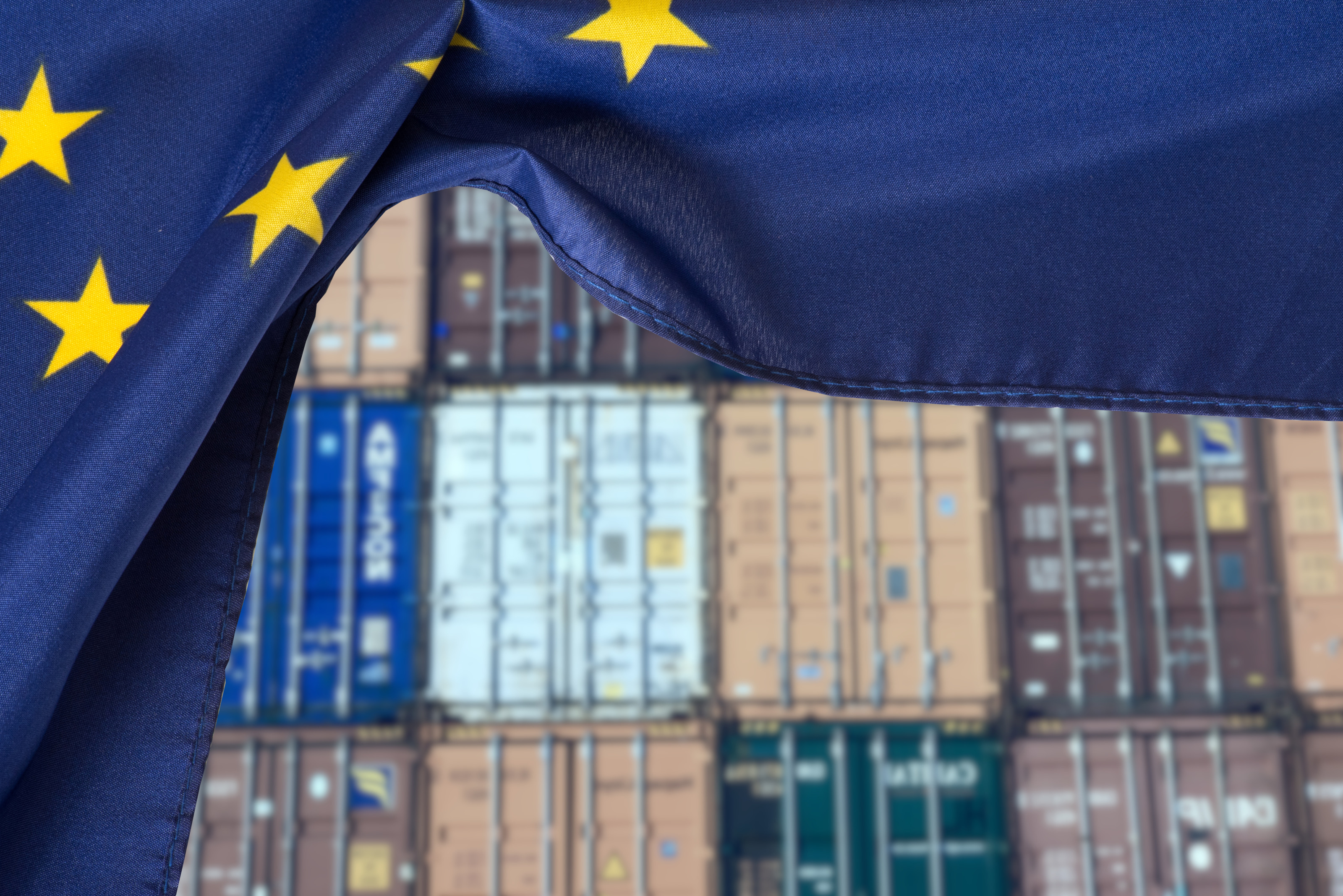 Im Vordergrund ist die Flagge der Europäischen Union, wie ein Vorhand zur Seite geschoben. Dahinter sind gestapelte Container, um Waren zu transportieren