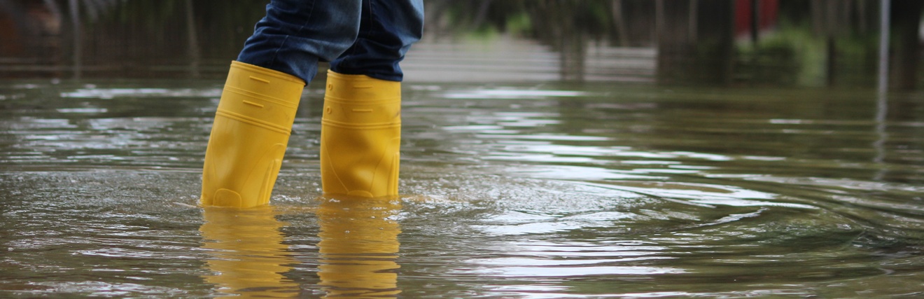 Person in Jeans steht mit gelben Gummistiefeln in tiefem Wasser, Hochwasser