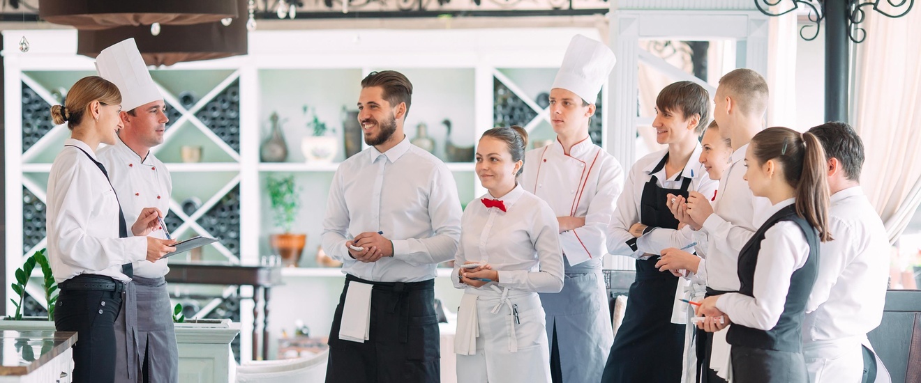 Mehrere Personen in Gastronomiegewändern, wie Schürzen und Kochhauben, stehen in Restaurant und blicken auf zwei weitere Personen ihnen gegenüber stehend