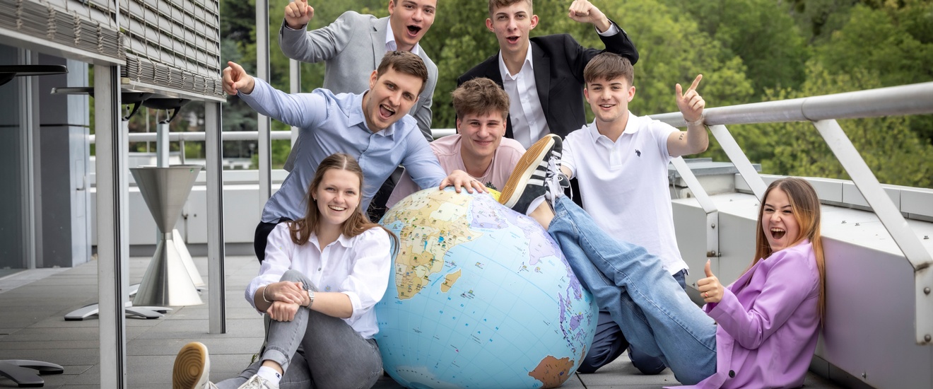 7 Lehrlinge sind um einen aufblasbaren Globus platziert und jubeln oder lächeln in Richtung Kamera.