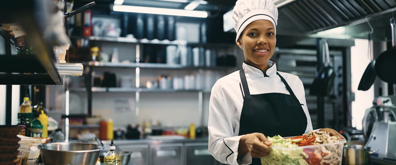 Eine Person mit Kochmütze, Kochkleidung und Schürze steht in einer Gastroküche und blickt freudig in die Kamera. In den Händen hält sie eine Plastikbox, befüllt mit geschnittener Zucchini, Paprika und Pilzen.
