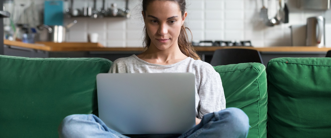 Person sitzt im Schneidersitz auf grünem Sofa und blickt auf aufgeklapptes Notebook am Schoß, im Hintergrund verschwommen Küchenzeile