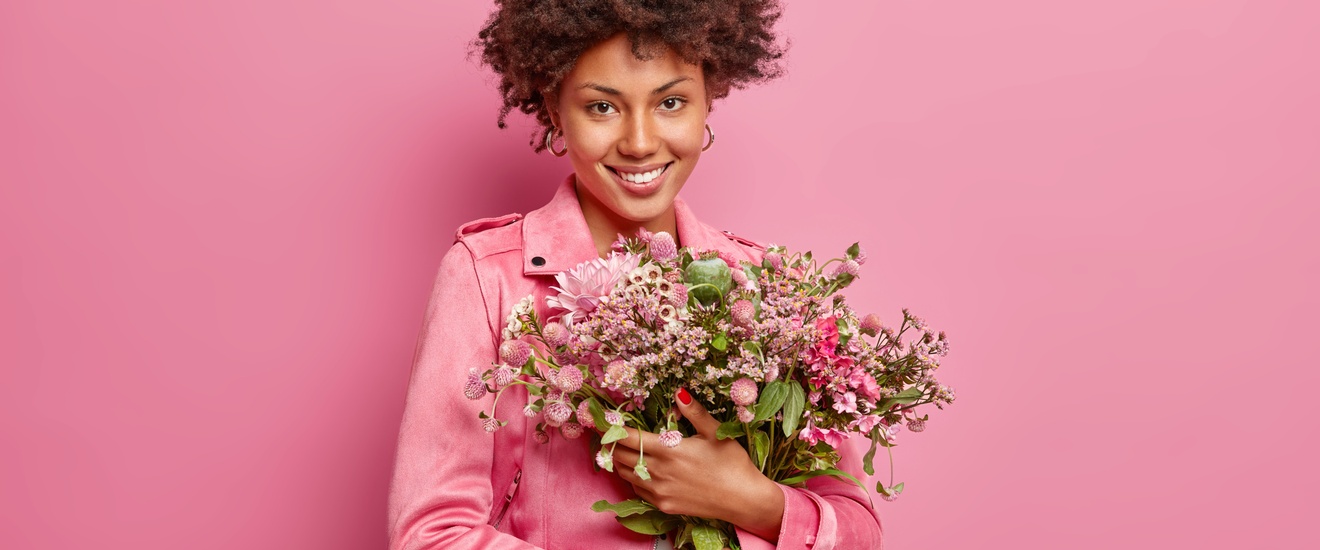 Lächelnde Person in pinker Jacke hält Blumenstrauß mit vorwiegend pinken Blüten, Hintergrund pink