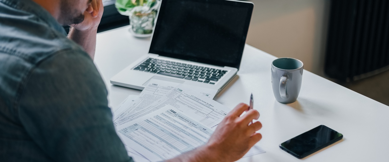 Person mit Brillen sitzt an Tisch und blickt auf Formulare vor ihr liegend, am Tisch aufgeklapptes Notebook, Tasse und Smartphone