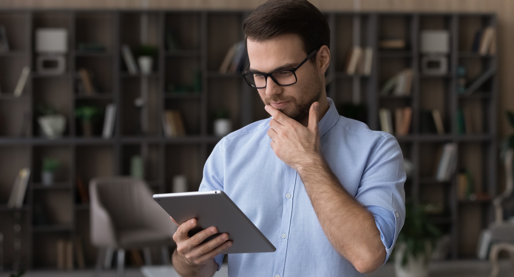 Person mit Brillen hält eine Hand an Kinn und blickt auf Tablet, im Hintergrund Bücherregal