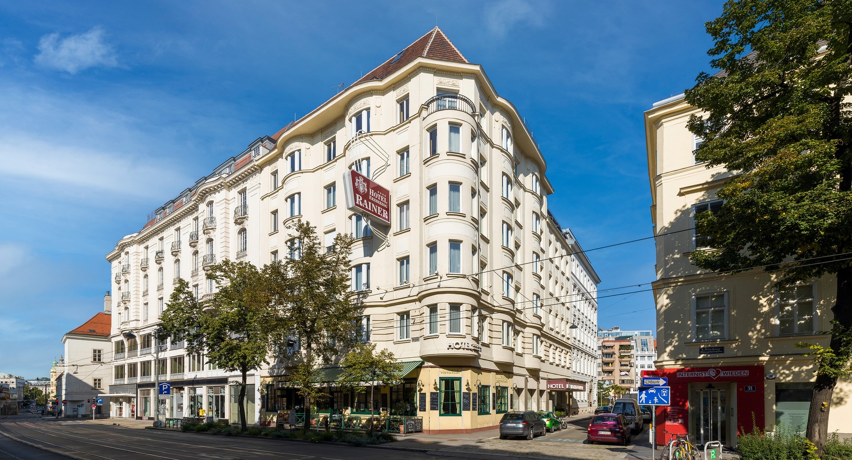 Außenansicht eines beigegelben historischen Eckgebäudes mit rotem Schild und weißem Schriftzug Hotel Erzherzog Rainer unter blauem Himmel
