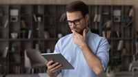 Person mit Brillen hält eine Hand an Kinn und blickt auf Tablet, im Hintergrund Bücherregal