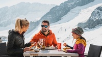 Ein Mann, eine Frau und ein Kind sitzen im Freien an einem Tisch und speisen. Im Hintergrund eine verscheite Berglandschaft.