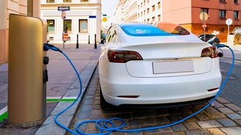 Weißes Elektroauto in Rückansicht, in dessen Stromladetank blauer Charger steckt verbunden mit Zapfsäule, im Hintergrund Gebäude einer Stadt