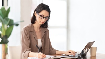 Person mit Brille sitzt an einem Tisch mit Laptop und einer Tasse und schreibt in ein Buch