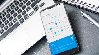 Smartphone mit geöffnetem digitalen Kalender liegt auf der Tastatur eines Laptops und eines dunklen Untergrunds, daneben ein angeschnittener Spiralblock sowie ein Kugelschreiber