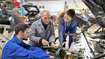 Zwei Lehrlinge arbeiten an Automotor, andere Person in grauem Mantel deutet auf Tablet