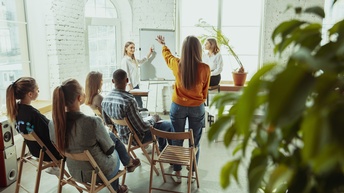 Personen sitzen und stehen in einem Raum mit Flipchart und tauschen sich aus