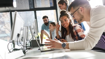 Personen sitzen und stehen rund um einen Schreibtisch und blicken dabei auf Laptops und Monitore vor ihnen