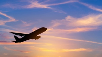 In der linken Bildhälfte ist die Silhouette eines Flugzeugs. Hinter dem Flugzeug ist ein bläulich-rötlicher Himmel mit vereinzelten Wolken bei Sonnenuntergang