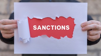 Nahaufnahme eines Zettels, auf dem Sanctions steht. Der Zettel wird von den Händen einer Person in die Kamera gehalten