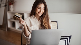 Person mit langen dunklen Haaren sitzt auf einem Sessel und blickt auf einen Laptop, der am Schoß platziert ist, mit der rechten Hand wird eine EC-Karte gehalten