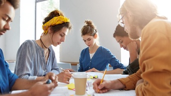 Jugendliche Personen sitzen gemeinsam bei einem Tisch, blicken auf Unterlagen und lernen