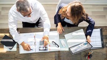 Zwei Personen in Businesskleidung halten ein Meeting an einem Schreibtisch mit einem Laptop und weiteren Arbeitsunterlagen wie Mappen, Taschenrechner, Listen, Notizbücher, Brillen und Stiften, Topview