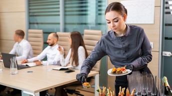 Servicepersonal in Arbeitskleidung mit schwarzen Handschuhen hält ein Teller mit Häppchen in der einen Hand und in der anderen Hand ein Häppchen mit Besteck von einem Buffet, im Hintergrund sitzen Personen in Businesskleidung bei einem Besprechungstisch