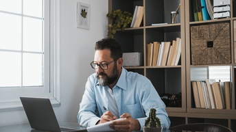 Person mit Brillen und Bart blickt auf aufgeklapptes Notebook und nimmt mit Stift auf Block Notizen vor, im Hintergrund Bücherregal