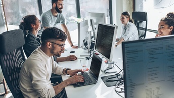 Personen sitzen freudig in einem Büro an einem Schreibtisch mit Laptop und Monitoren