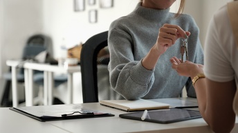 Person übergibt einen Schlüssel an eine andere Person, am Tisch liegen diverse Unterlagen