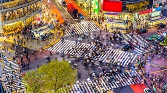 Topshot einer großen Kreuzung mit vielen Zebrastreifen, auf denen sehr viele Menschen gehen. Neben den Menschen stehen Fahrzeuge auf den Straßen. An den Straßen sind beleuchtete Gebäude