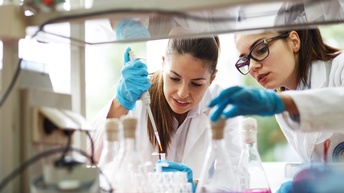 Zwei Personen mit blauen Gummihandschuhen in Labor, eine Person füllt mit Füllstab Flüssigkeit in Eprouvette, andere Person greift nach Reagenzglas mit violetter Flüssigkeit