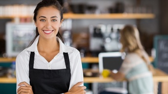 Person mit dunklen geschlossenen Haaren, weißer Bluse und Arbeitsschürze steht in einem Lokal vor einer Theke, im Hintergrund sitzt eine Person mit einem Laptop bei einem Tisch