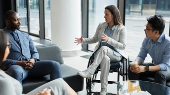 Personen in Businesskleidung unterhalten sich in einer Besprechungszone, eine Person sitzt dabei in einem Rollstuhl und teilt etwas mit