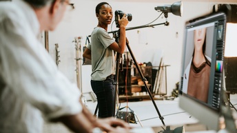 Person hinter Fotokamera in Fotostudio stehend blickt zu anderer Person verschwommen im Vordergrund, die vor Computermonitor mit Bildbearbeitungsprogramm steht