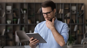 Person mit Brillen hält eine Hand an Kinn und blickt auf Tablet, im Hintergrund Bücherregal