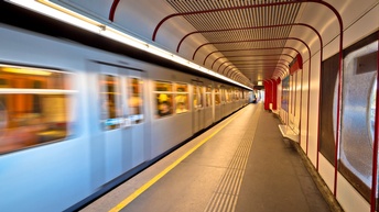 In der linken Bildhälfte sind mehrere silberne Waggons einer sich bewegenden U-Bahn. Rechts daneben ist ein asphaltierter Boden mit an den Wänden montierten Bänken