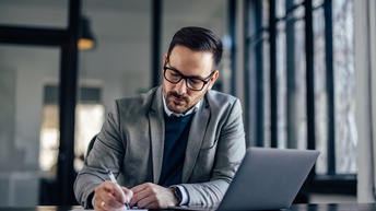 Person mit kurzen Haaren, Brille und Bart trägt Businesskleidung und arbeitet bei einem Schreibtisch mit einem Laptop in einem Büro