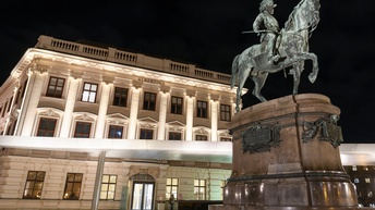 Außenaufnahme eines großen Gebäudes vor dunklem Himmel, auf dem Albertina steht. Hinter ein paar Fenstern ist Licht. Davor steht eine Statue mit einem Pferd und einem Reiter auf einem Sockel.