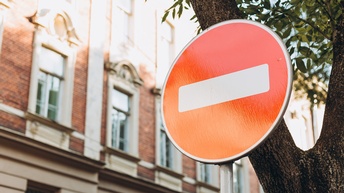 Detailansicht eines Straßenschildes für Fahrverbot: Kreisrundes rotes Schild mit weißem Streifen mittig, im Hintergrund Ast eines Baumes und verschwommen Hausfassade