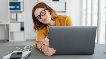 Person mit rötlichen, schulterlangen Haaren, Brille sowie gelber Bluse sitzt freudig bei einem Schreibtisch und hält den aufgeklappten Laptop, am Tisch liegen Notizbuch, Smartphone, Kopfhörer und im Hintergrund zeigen sich weiße Regale mit Ordnern