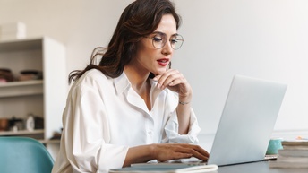 Seitliche Aufnahme einer an einem Tisch sitzenden Person mit Brille, roten Lippen und heller Bluse. Vor ihr steht ein aufgeklappter Laptop, auf dem sie ihre rechte Hand hat. Ihre linke Hand ist unter ihrem Kinn