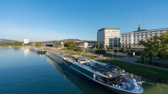 Schiff liegt an Ankerplatz auf Fluss an sonnigem Tag mit blauem Himmel, daneben Gebäude einer Stadt und Grünflächen