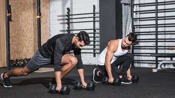 Zwei bärtige Personen bei Dehnungsübungen mit Hanteln am Boden eines Raumes für Fitnessübungen, im Hintergrund weitere Sportgeräte und Gewichte