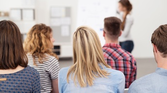 Rückenansicht mehrerer sitzender Personen, eine Person vorne bei Flipchart stehend