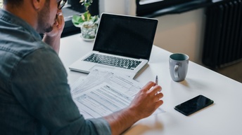 Person mit Brillen sitzt an Tisch und blickt auf Formulare vor ihr liegend, am Tisch aufgeklapptes Notebook, Tasse und Smartphone