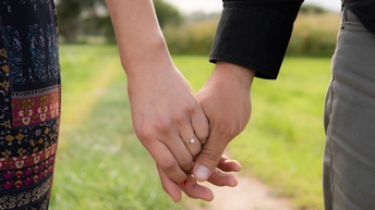 Detailaufnahme von zwei sich haltenden Händen zweier Personen,  eine Hand trägt einen Ring mit funkelndem Stein, dahinter zeigt sich ein Weg in einer Naturlandschaft