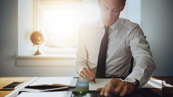 Person in Businesskleidung sitzt an einem Schreibtisch und markiert ein Dokument während mit einem Taschenrechner gerechnet wird, daneben liegt ein Smartphone sowie ein Notizbuch und im Hintergrund steht ein kleiner Globus