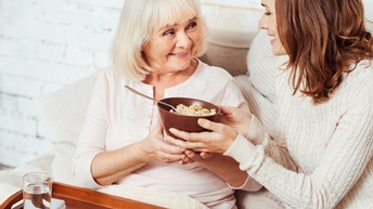 Person reicht einer älteren, pensionierten und strahlenden Person, die auf einer Couch sitzt, ein Müsli während ein Glas Wasser und Tabletten auf einem Tablet stehen