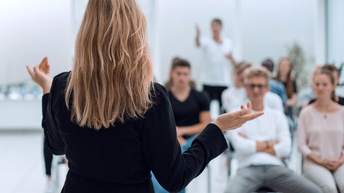 Rückansicht einer sprechende Person vor lauschenden Zuhörer:innen