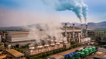 Fabriksgelände mit Silos und Schornsteinen aus denen Rauch austritt, im Hintergrund hügelige Landschaft