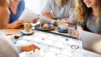 Mehrere lächelnde Personen sitzen um Tisch und blicken auf darauf ausgebreiteten Terminkalender