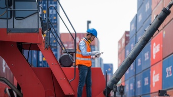 Person mit roter Warnweste und blauem Schutzhelm hält ein Tablet in Händen und steht auf einem Containerstapler, im Hintergrund zeigen sich viele gestapelte Container